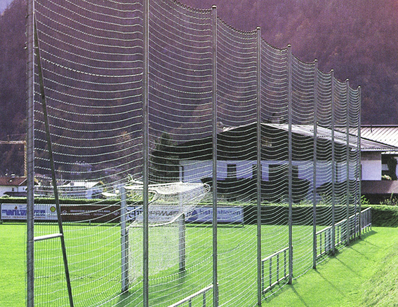 Ein robustes Schutznetz hinter einem Fußballtor auf einem grünen Sportplatz mit umliegenden Gebäuden im Hintergrund.