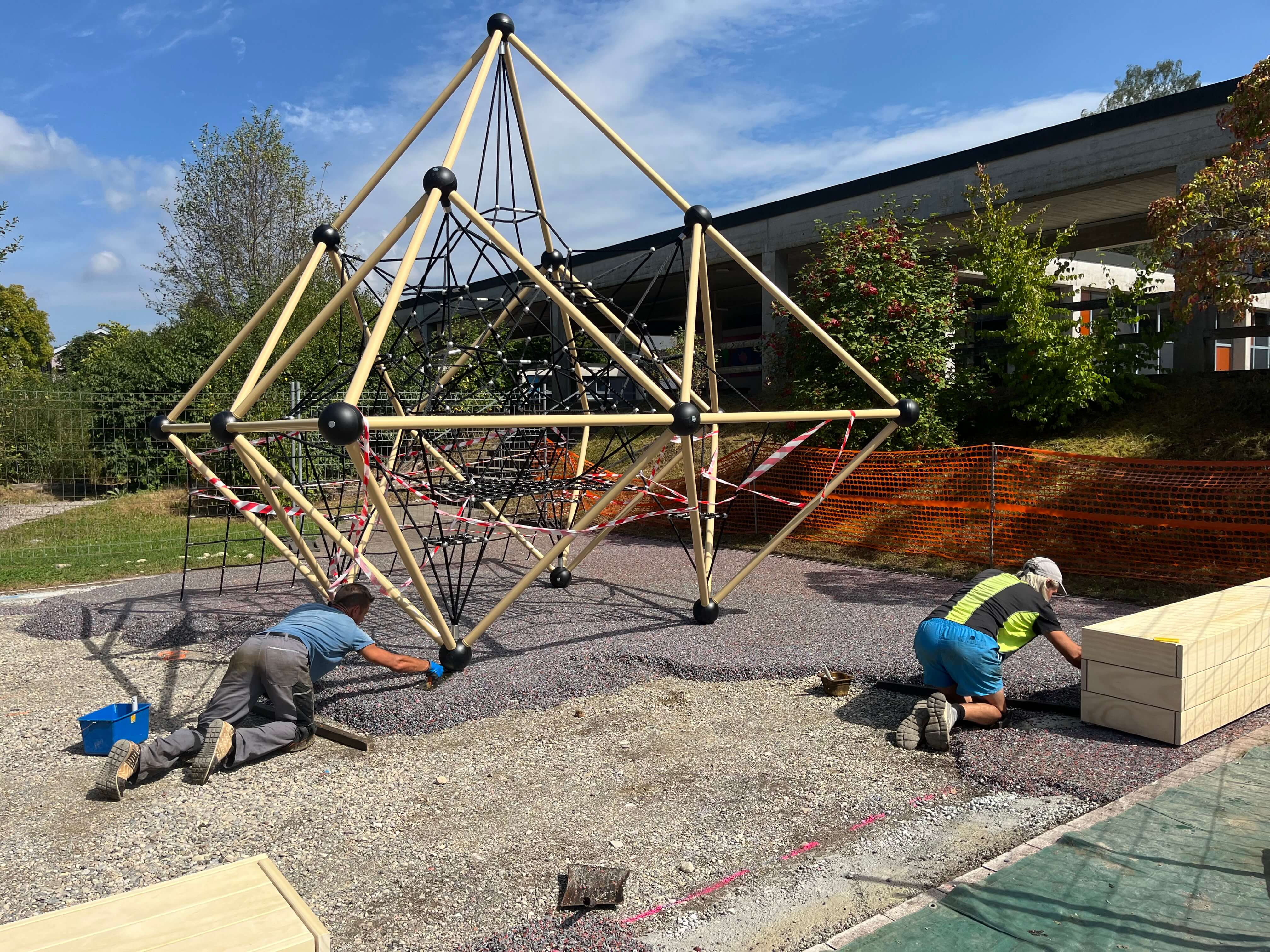 Zwei Männer installieren ein pyramidenförmiges Klettergerüst aus Holz und Seilen auf einem Spielplatz. Im Hintergrund sind Bäume und ein Gebäude zu sehen.