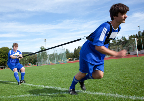 Zwei Fußballspieler in blauen Trikots beim Widerstandstraining auf einem Sportplatz, einer zieht den anderen mit einem Gummiseil.