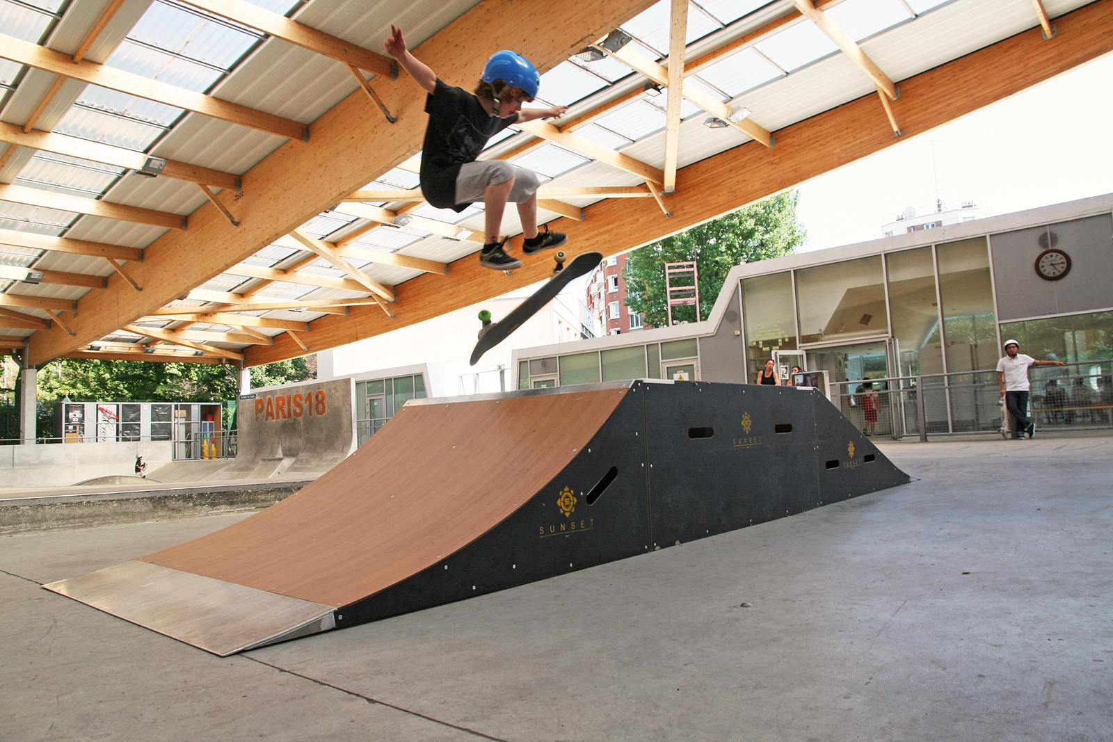 Kind mit Helm macht einen Skateboard-Trick über eine Rampe in einem überdachten Skatepark