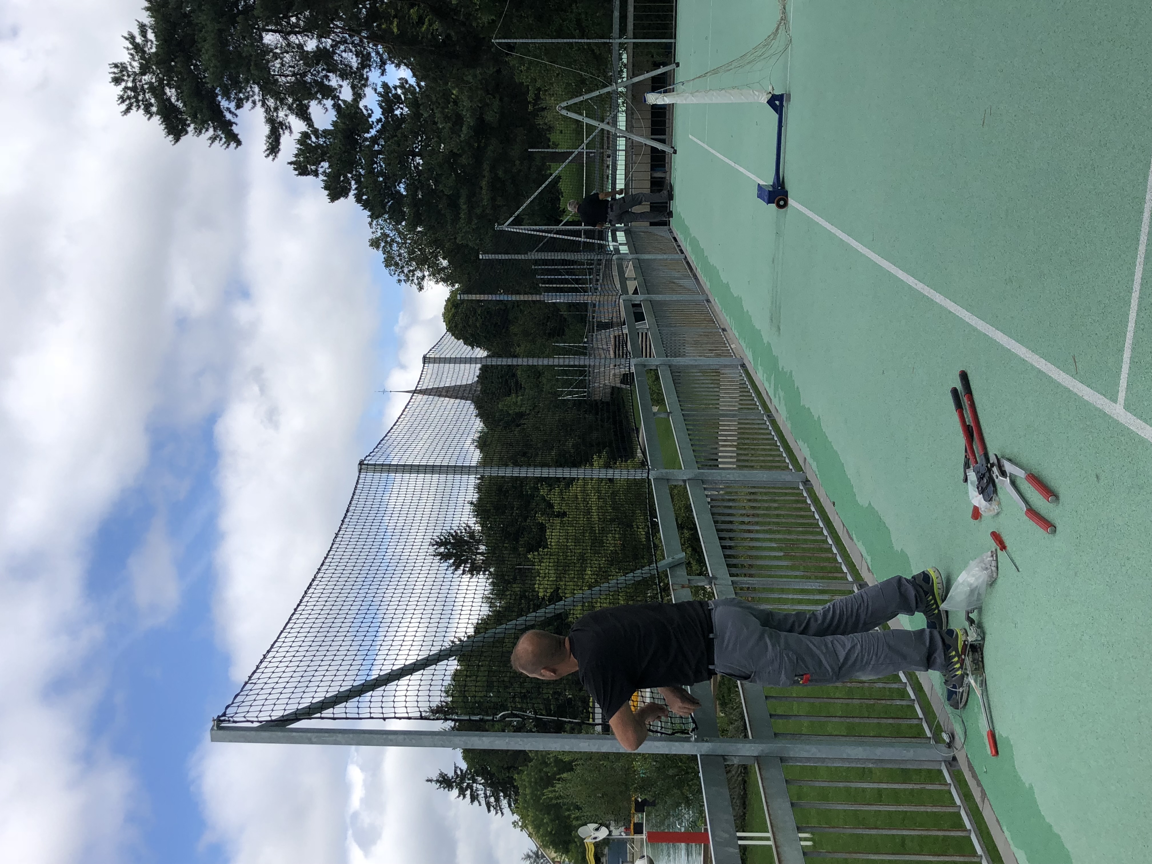 Zwei Männer installieren ein Sicherheitsnetz auf einem Sportplatz. Im Vordergrund liegt Werkzeug auf dem Boden, und im Hintergrund sind Bäume und ein Kirchturm zu sehen.