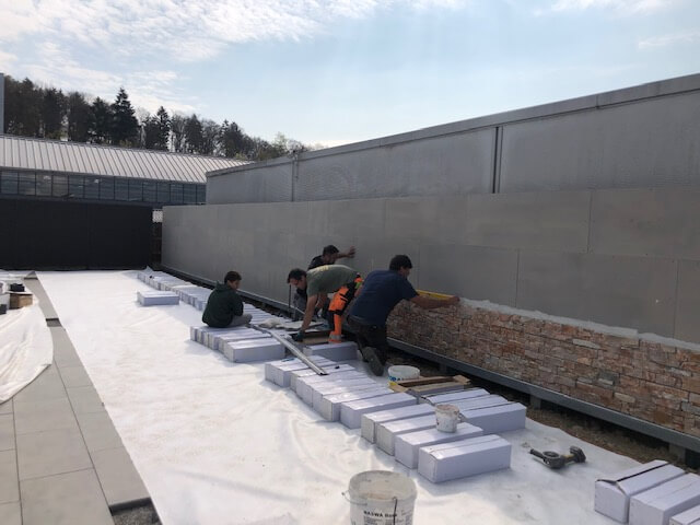 Mehrere Arbeiter verlegen Fliesen auf einer großen Baustelle im Freien an einer Wand.