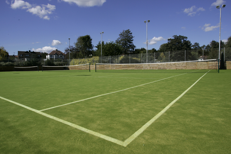 Ein leerer Tennisplatz mit grünem Kunstrasen unter blauem Himmel und einigen Wolken, umgeben von Bäumen und Zäunen.