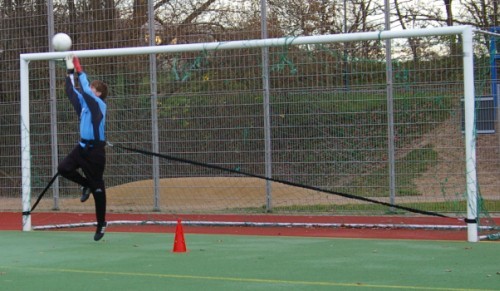 Ein Torwart in blauer Kleidung springt hoch, um einen Fußball am Tor zu blockieren, mit einem roten Trainingskegel im Vordergrund auf einem Fußballfeld