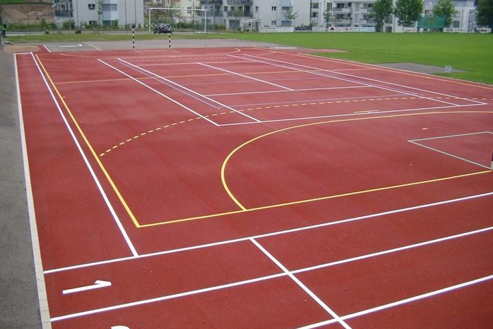 Großes, rotes Sportfeld mit Markierungen für verschiedene Spiele wie Basketball, Tennis und Fußball auf einem Sportplatz.
