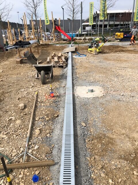 Baustelle mit einer installierten Entwässerungsrinne, umgeben von Baumaterialien und Werkzeugen, mit einem neu entstehenden Spielplatz im Hintergrund