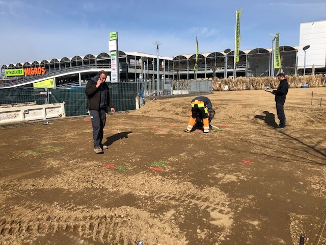 Drei Personen bei der Arbeit auf einer Baustelle vor einem Einkaufszentrum bei strahlend blauem Himmel