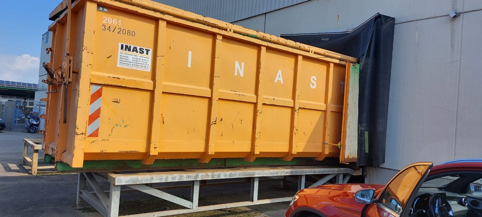 Ein orangefarbener Müllcontainer mit der Aufschrift 'INAST', auf einem Metallgestell an ein Gebäude gelehnt.