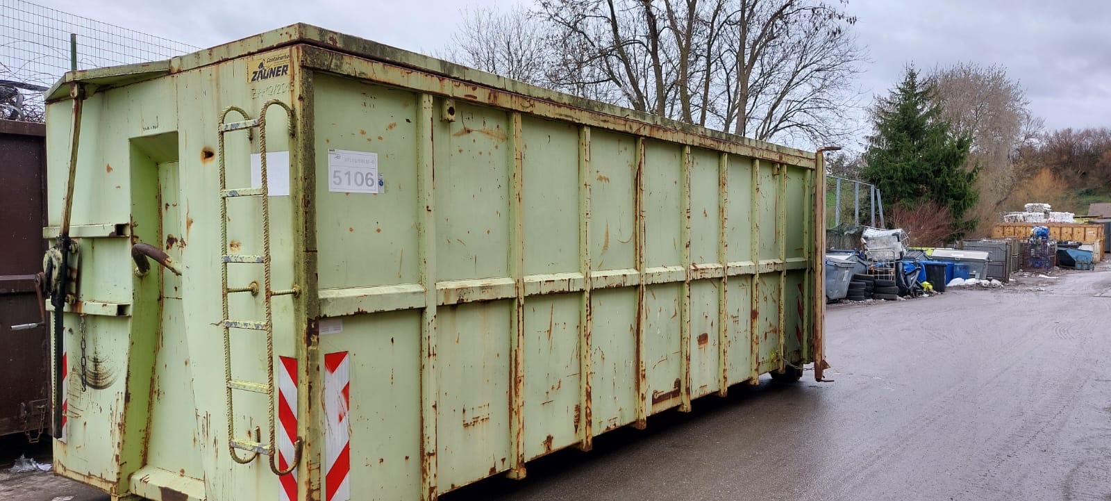 Ein großer, grüner Sperrmüllcontainer steht auf einem Recyclinghof. Im Hintergrund sind Bäume und weitere Container zu sehen.