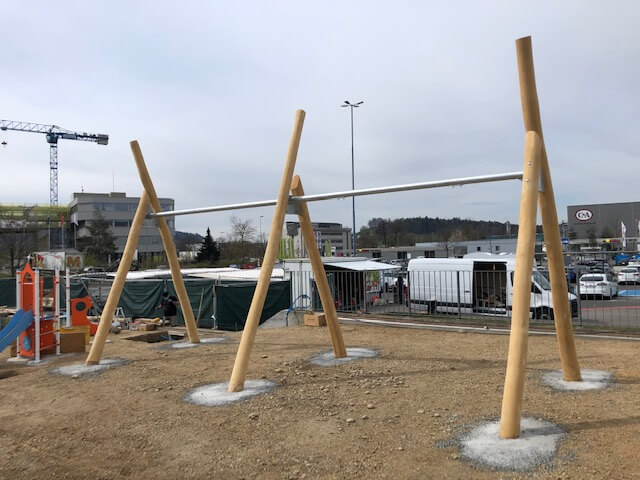 Unfertige Spielplatzkonstruktion aus Holz mit schräg stehenden Balken, aufgenommen auf einer Baustelle vor einem Industriegebiet