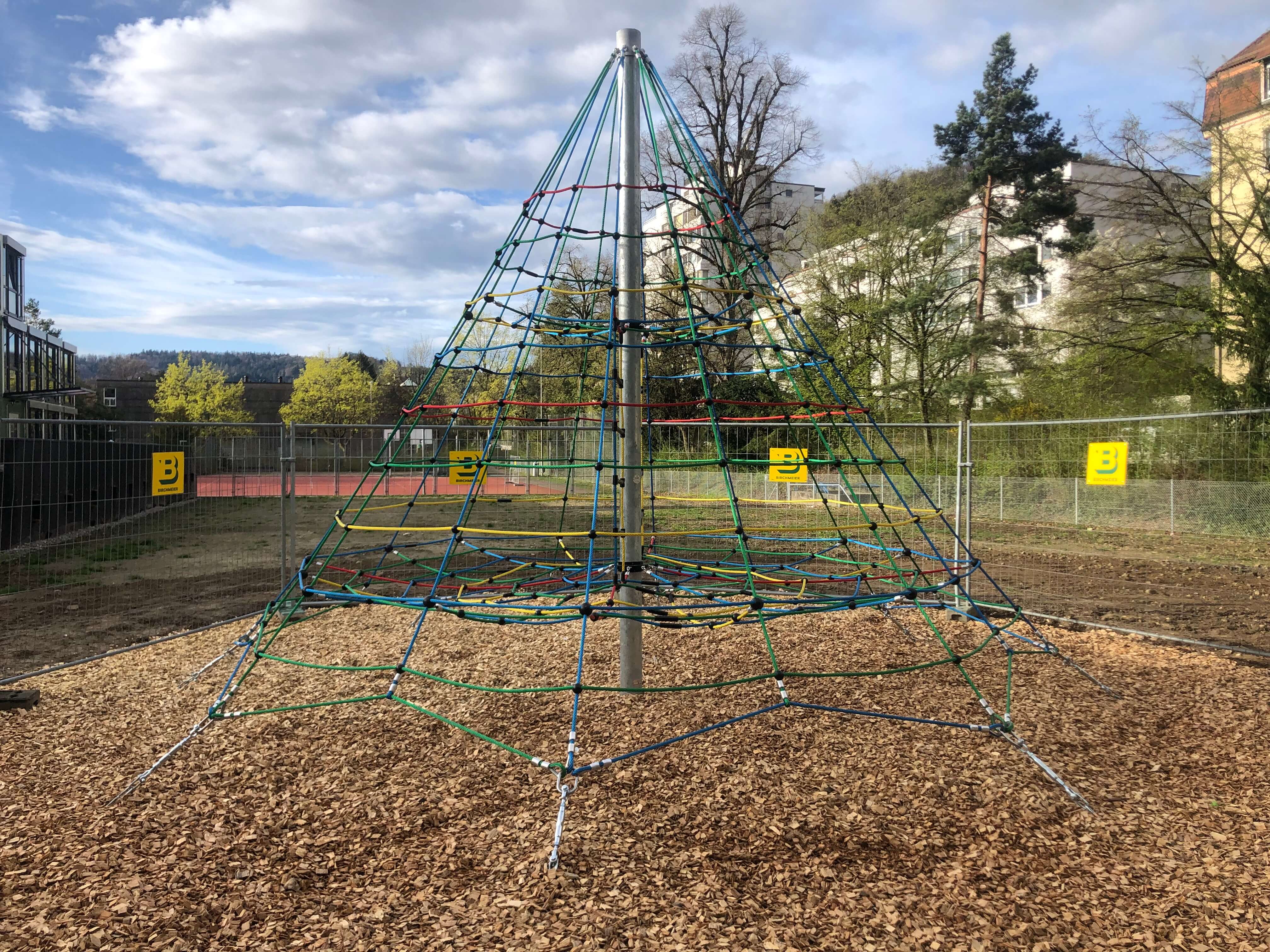 Bunte Kletterpyramide aus Seilen, umgeben von Zaun mit Holzhackschnitzeln als Bodenbelag auf einem Spielplatz im Freien