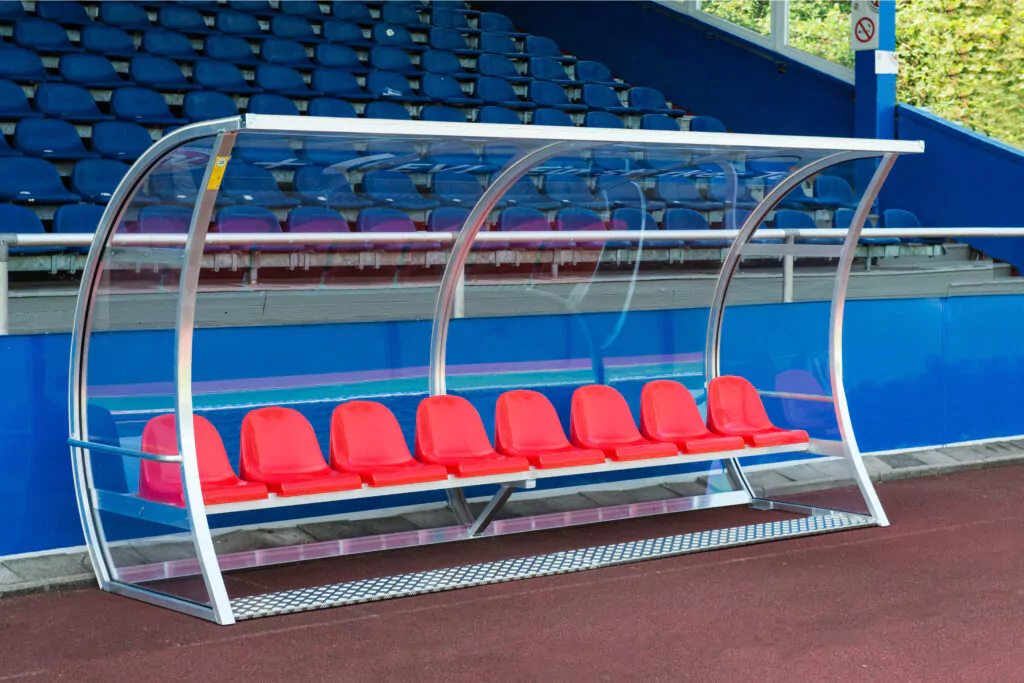 Ersatzbank mit roten Kunststoffsitzen unter einem transparenten Überdach im Stadion