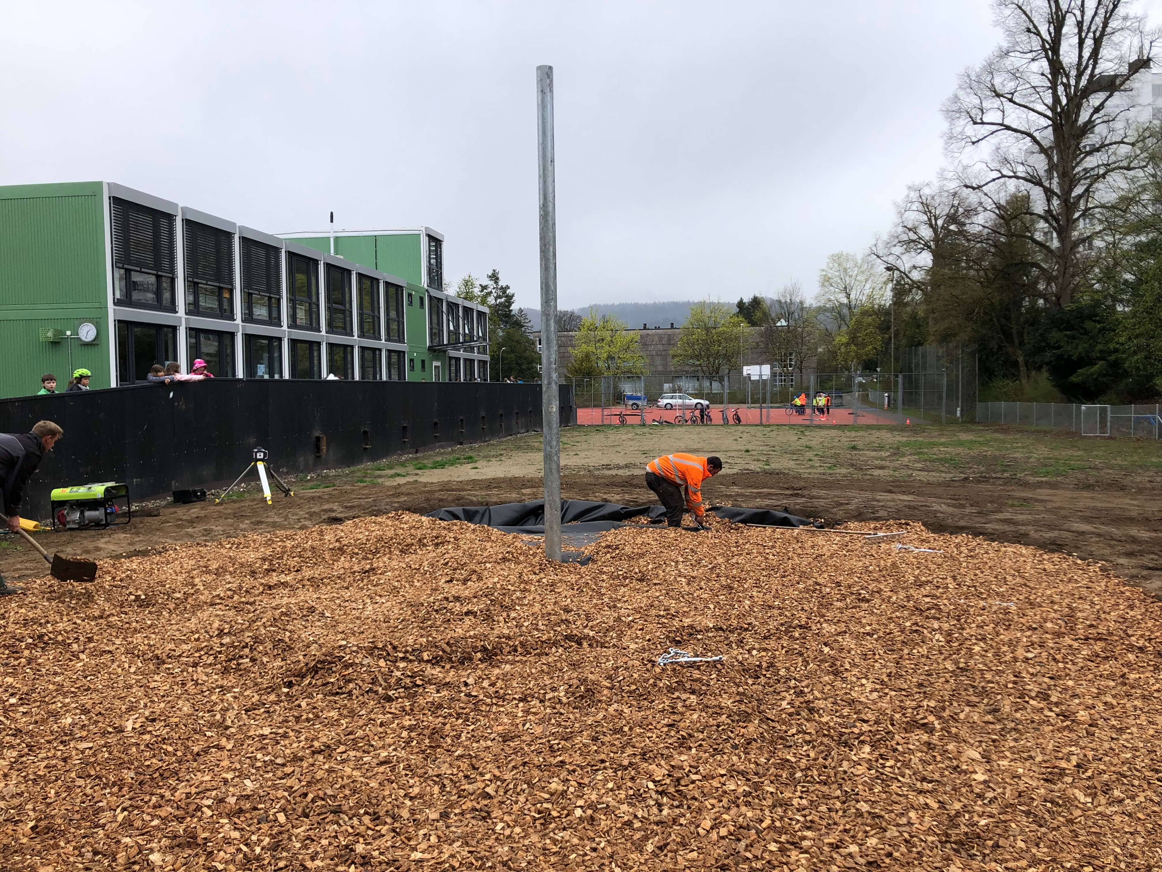 Eine Baustelle mit Holzschnitzeln und zwei Bauarbeitern vor einem Gebäude mit grüner Fassade