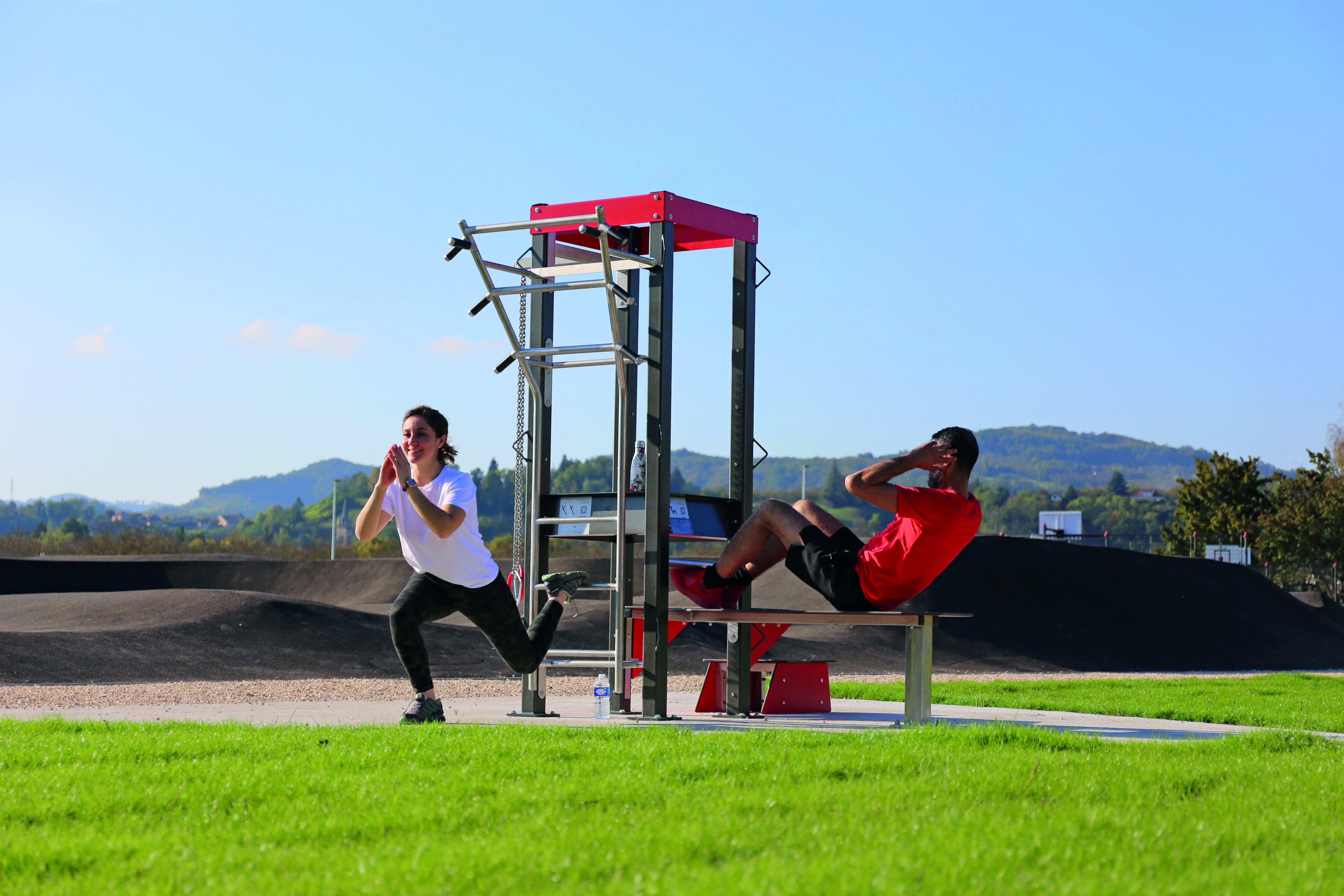 Zwei Personen machen Sportübungen an einem Outdoor-Fitnessgerät auf einer Wiese