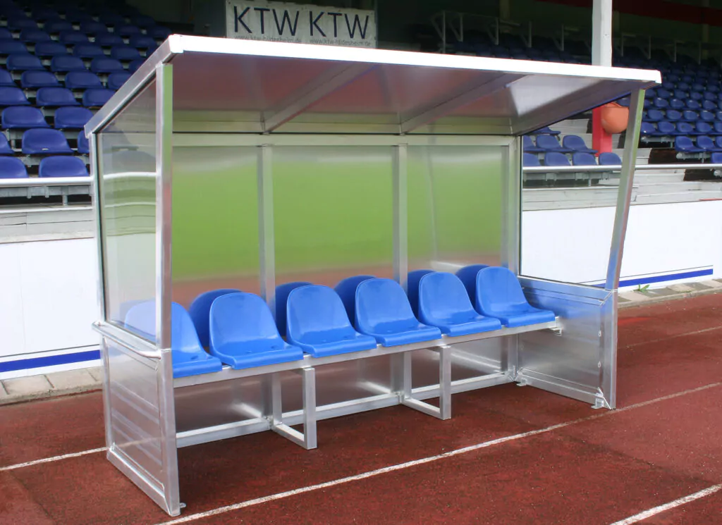 Überdachte Fußball-Ersatzbank mit mehreren blauen Sitzen auf einem Sportplatz