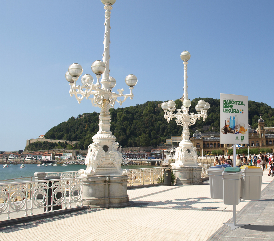 Elegante weiße Laternen stehen auf einer Promenade am Meer mit Blick auf die Stadt und bewaldete Hügel im Hintergrund.