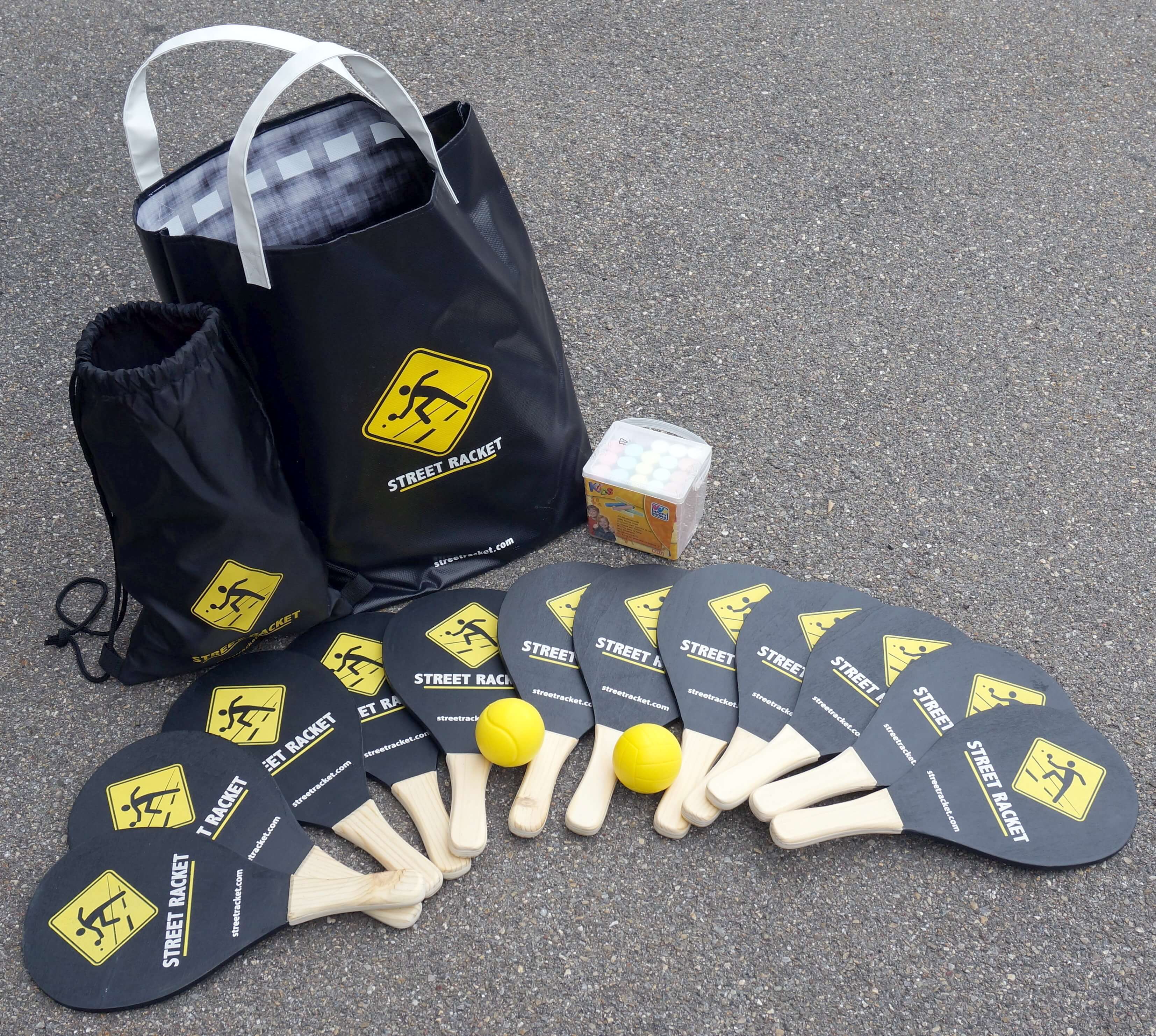 Street Racket Spielset mit mehreren Schlägern, gelben Bällen, einem Würfelset und zwei schwarzen Taschen auf Asphalt