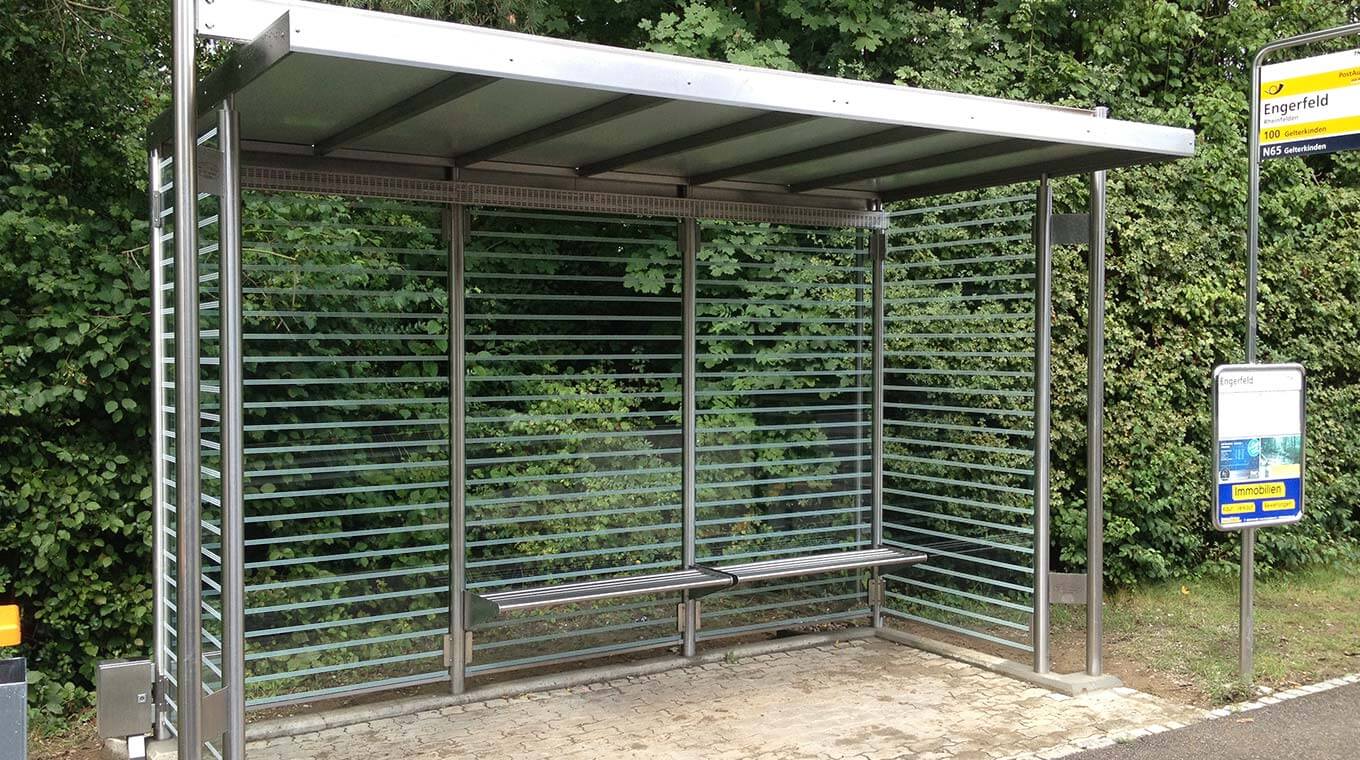 Bushaltestelle mit Glaswänden, metallener Sitzbank und Überdachung in einer grünen Umgebung