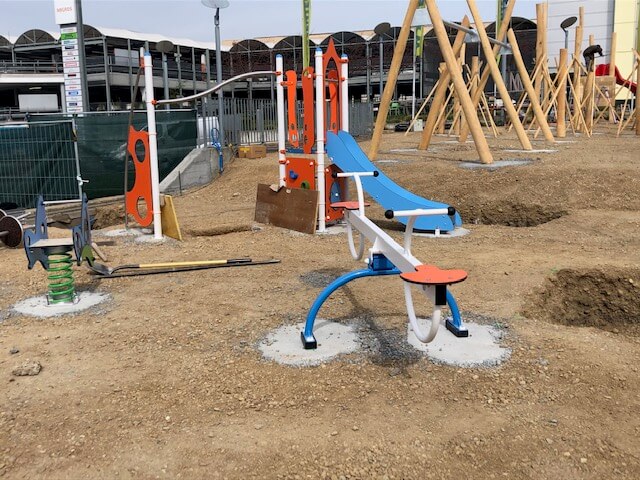 Ein unfertiger Spielplatz mit farbenfroher Wippe und Rutsche, umgeben von Baustellenmaterialien.