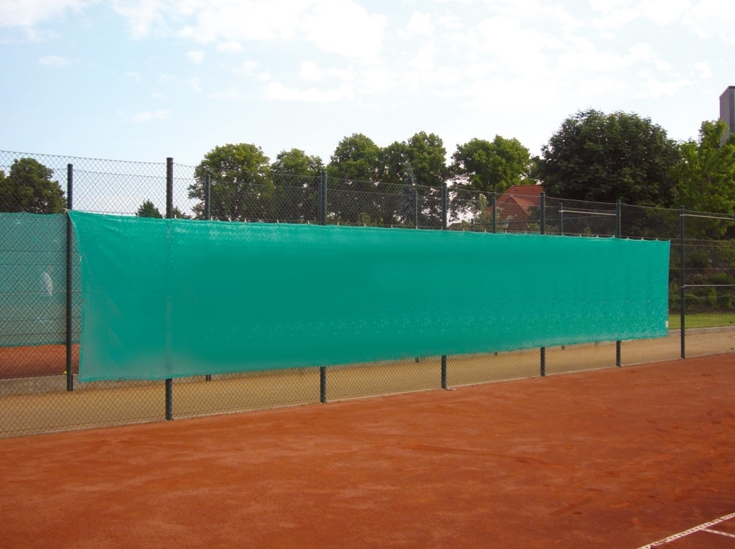 Grüne Sichtblende auf einem Tennisplatz mit rotem Sandboden und umgebender Vegetation