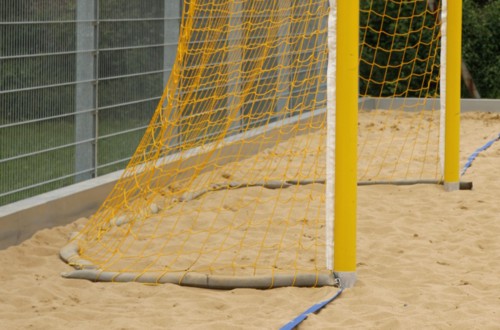 Gelbes Strandfußball-Tor mit Netz auf einem Sandplatz, seitlich von einem Metallzaun umgeben