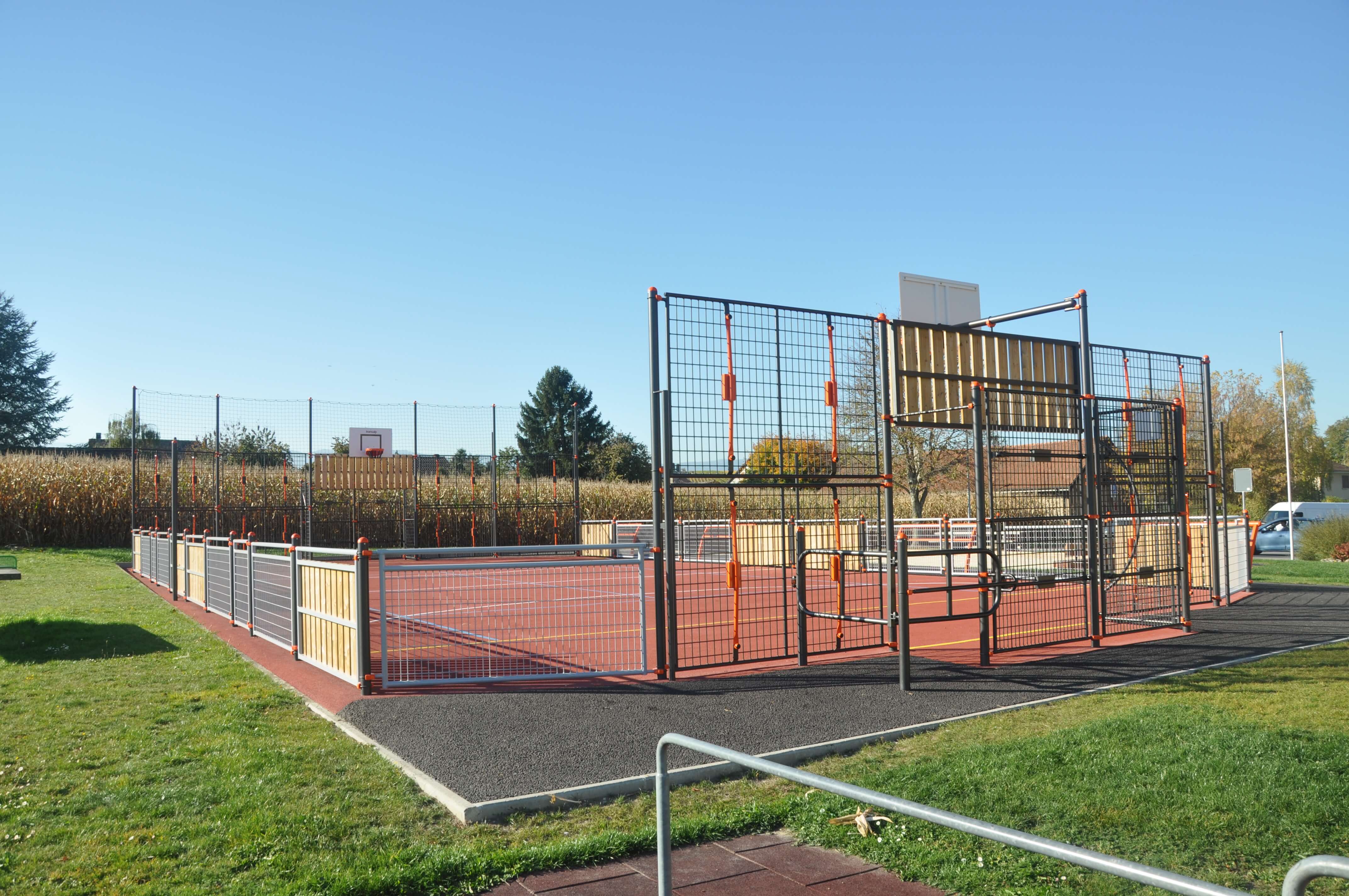 Ein moderner Basketballplatz mit Schutzzaun, umgeben von grünem Rasen und Bäumen, an einem sonnigen Tag