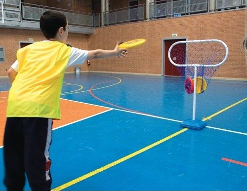 Junge im gelben Trikot wirft eine Frisbee-Scheibe in ein freistehendes Netz in einer Turnhalle.