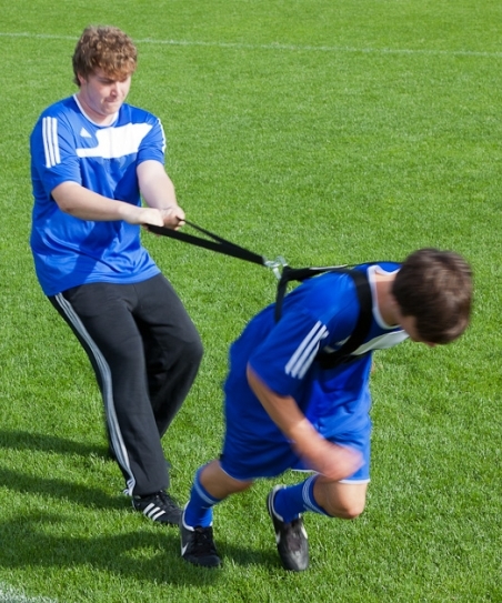 Zwei junge Männer beim Fußballtraining, wobei einer den anderen mit Widerstandsbändern auf einem Sportplatz zurückhält.