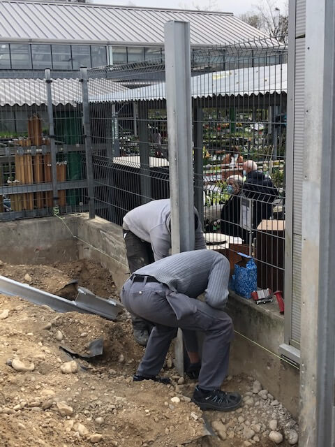 Zwei Arbeiter errichten einen Metallzaun auf einer Baustelle, während im Hintergrund eine Pflanzenausstellung zu sehen ist.