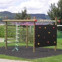 Ein Spielplatz-Klettergerüst mit einer Boulderwand und einem Kletterseil, umgeben von grüner Landschaft und Bäumen im Hintergrund.