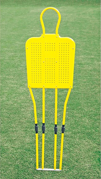 Ein gelber Fußball-Trainingsdummy steht auf einem grünen Rasenfeld. Der Dummy hat einen Rundbogen oben und besteht aus mehreren Stangen.