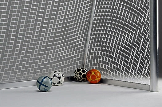 Vier unterschiedlich farbige Fußbälle liegen in einer Ecke eines leeren Fußballtors mit einem weißen Netz.