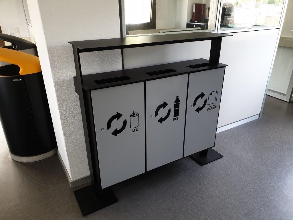 Graue Recyclingstation mit Symbolen zur Mülltrennung für Alu, PET-Flaschen und Papier in einem Innenbereich