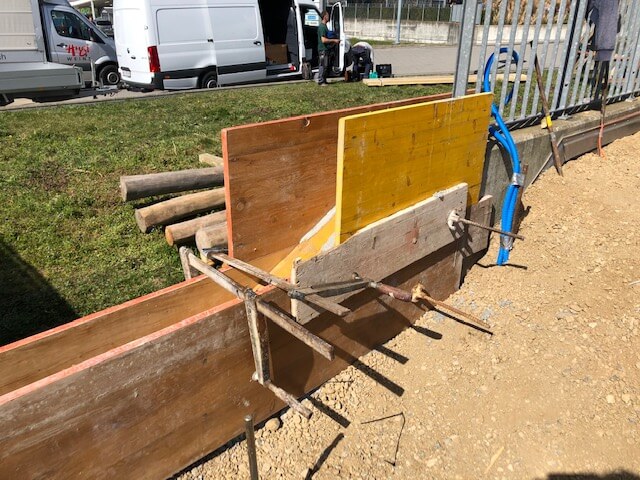 Holzschalungswände für Betonarbeiten auf einer Baustelle, mit im Hintergrund geparkten Lieferfahrzeugen und Rasenfläche.