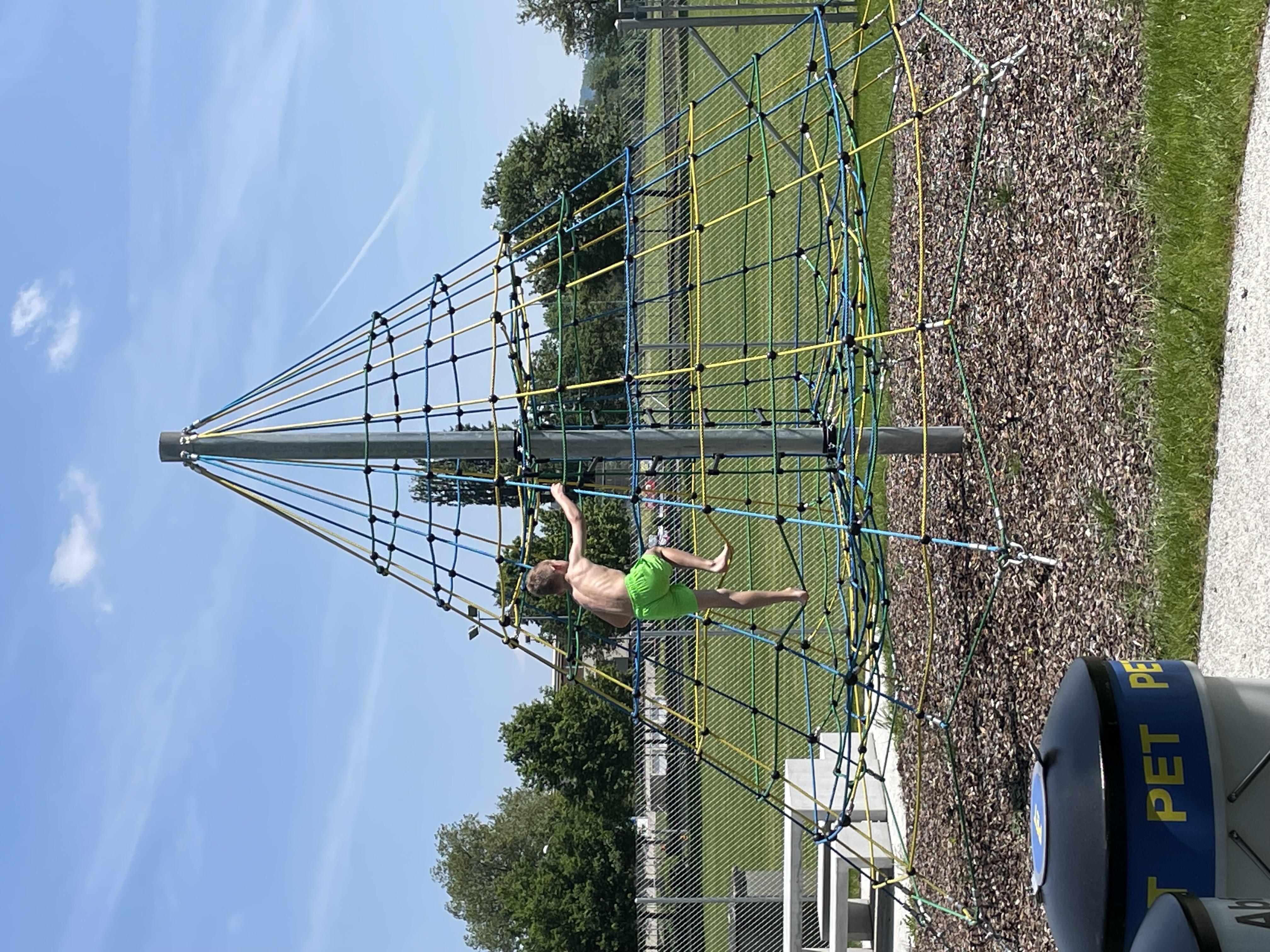 Ein Kind klettert auf einem pyramidenförmigen Klettergerüst auf einem Spielplatz bei sonnigem Wetter.