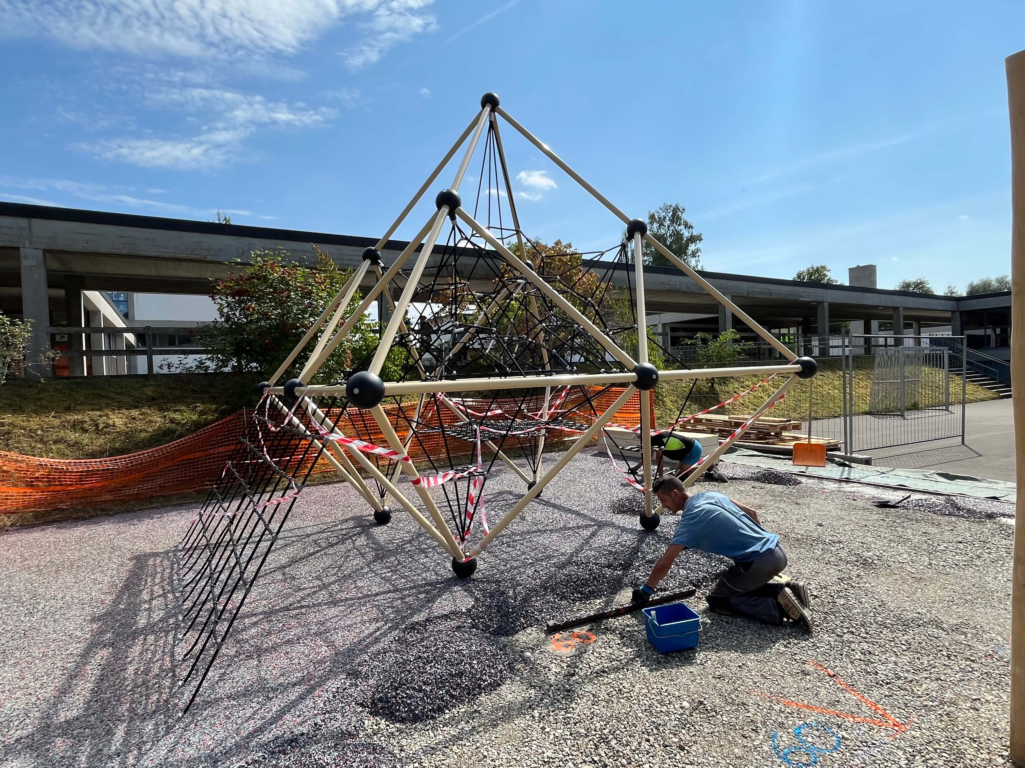 Ein Klettergerüst wird auf einem Schulhof montiert, während ein Arbeiter den Boden vorbereitet.