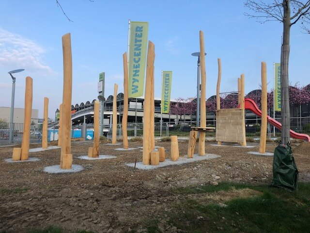Moderner Spielplatz mit großen Holzpfosten im Freien, umgeben von grüner Landschaft und blauen Himmel, mit einer Rutsche im Hintergrund
