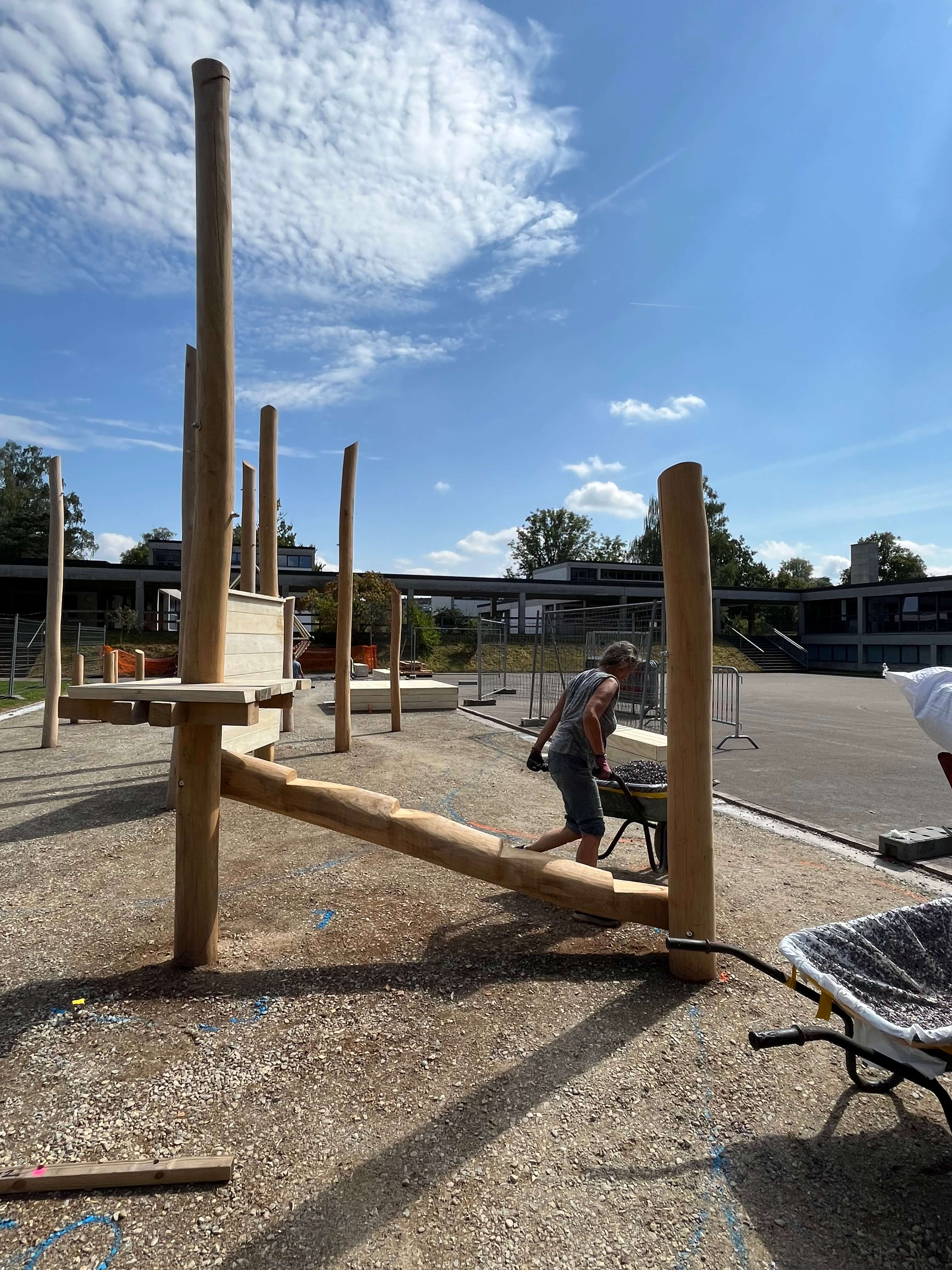 Ein Spielplatz mit einem großen hölzernen Klettergerüst im Bau, Arbeiter verwenden eine Schubkarre.