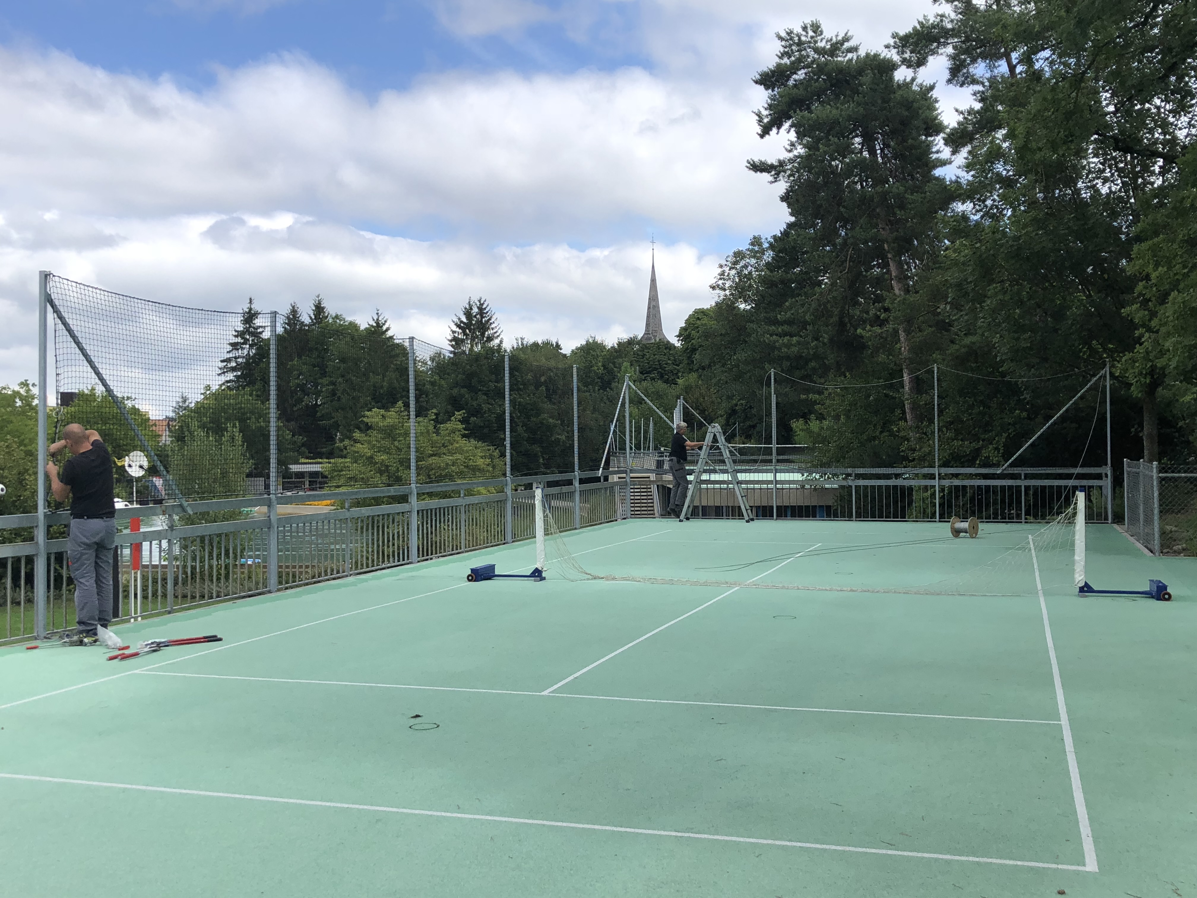 Zwei Menschen arbeiten an einem Netz auf einem Outdoor-Tennisplatz, umgeben von Bäumen und einem Kirchturm im Hintergrund.