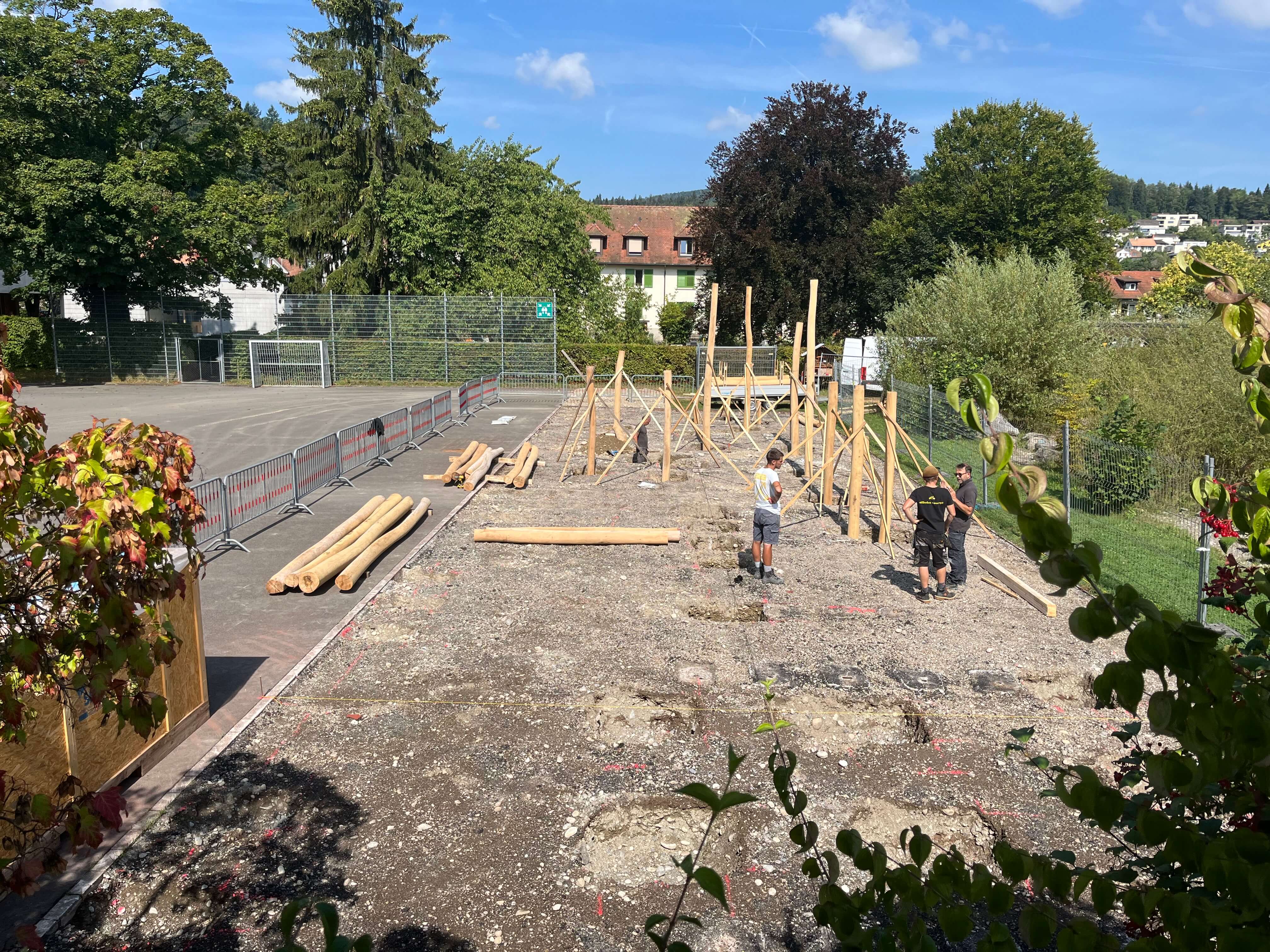 Arbeiter errichten ein hölzernes Klettergerüst auf einem Schulhof, umgeben von Bäumen und einem Fußballfeld.