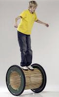 Kind im gelben T-Shirt balanciert auf einem großen Holz-Balance-Rad gegen einen neutralen Hintergrund