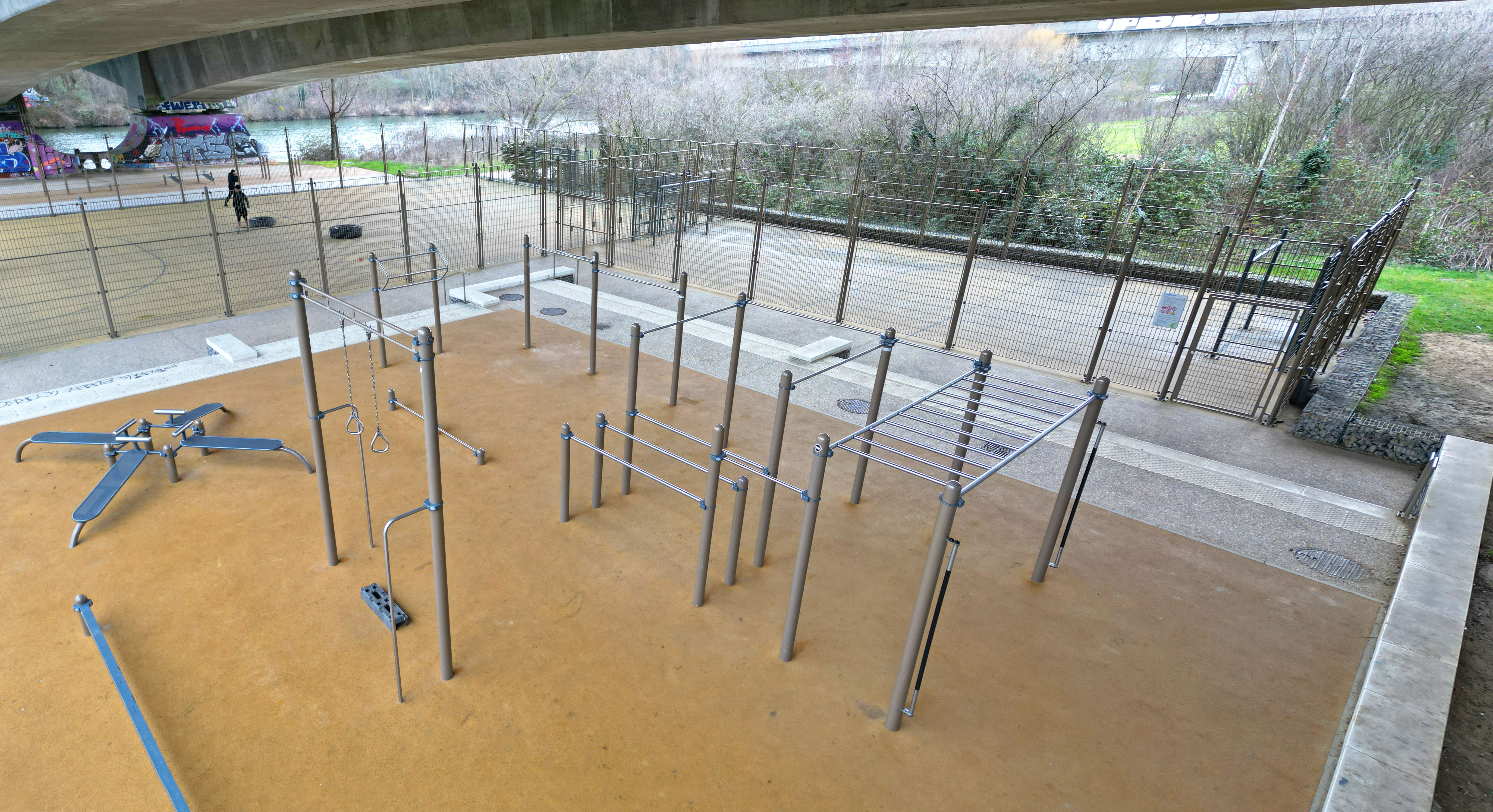 Ein moderner Outdoor-Fitnesspark unter einer Brücke mit verschiedenen Calisthenics-Geräten, darunter Klettergerüste und Trainingsstangen.